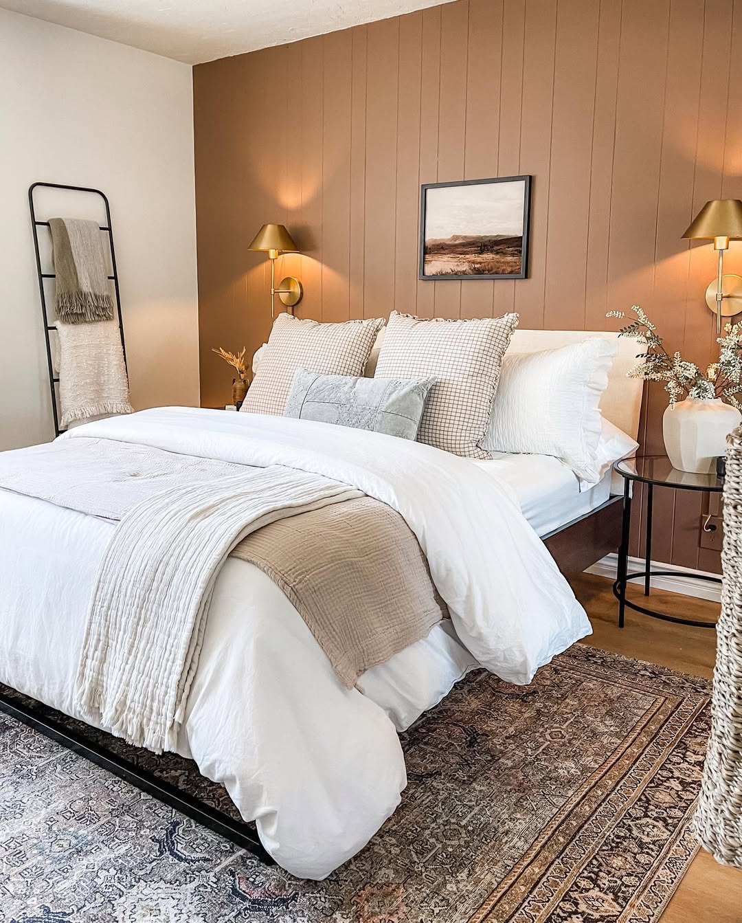 A cozy small guest bedroom with a neatly made bed adorned in white and beige linens, pillows, and a throw. A brown accent wall showcases a framed picture and wall sconces, while a ladder shelf holds a blanket and charming decor items.